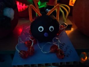 Zdjęcie czarnej futrzanej piłeczki z przyczepionymi oczkami i fioletowym nosem. Na czubku piłeczka zamontowane ma pomarańczowe uszka a nogi imitują dużę pomarańczowe lampki na białych kabelkach.