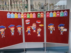 Zdjęcie dekoracji przygotowanej z okazji pasowania na ucznia. Na czerwonym tle kolorowy napis :"Pasowanie na ucznia" . Pod napisem wycięte z papieru podobizny usmiechniętychdzieci z torbami, przyborami szkolnymi, biretamina głowach i dyplomami oraz książki.