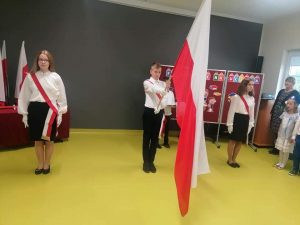 Zdjęcie pocztu flagowego podczas uroczystości pasowania na ucznia. Po bokach dwie uczennice stojące na baczność, a po środku uczeń trzymający częściowo opuszczoną flagę Polski.