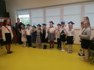 Uczniowie klasy pierwszej podczas pasowania na ucznia. Dzieci stoją obok siebie w strojach galowych z podniesionymi prawymi dłońmi w górę. Na głowach maja birety.