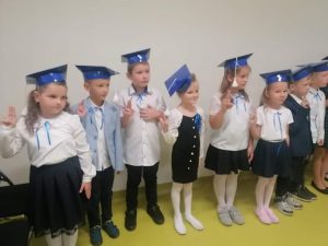 Uczniowie klasy pierwszej podczas pasowania na ucznia. Dzieci stoją obok siebie w strojach galowych z podniesionymi prawymi dłońmi w górę. Na głowach maja birety.