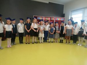 Uczniowie klasy pierwszej stoją w jednym rządku na sali teatralnej. Dzieci ubrane są w stroje galowe a na głowach mają birety.