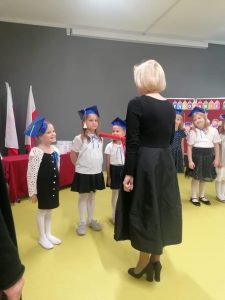 Pani Dyrektor pasuje dużą czerwoną kredką uczennicę kładąc jej ją na ramieniu. Obok stoją kolejne dzieci czekające na pasowanie.