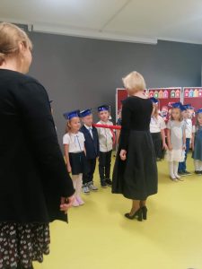 Pani Dyrektor pasuje dużą czerwoną kredką ucznia kładąc mu ją na ramieniu. Obok stoją kolejne dzieci czekające na pasowanie.