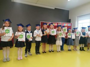 Uczniowie klasy pierwszej stoją w galowych strojach na sali teatralnej. Dzieci stoja w jednym rzędzie, na głowach mają birety a w dłoniach otrzymane pamiatkowe dyplomy i książeczki.