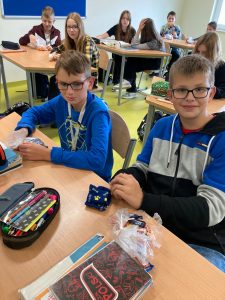 Uczniowie klasy siódmej pozują do zdjęcia. Przed nimi leżą złożone skarpetki - prezent z okazji dnia chłopaka. W tle widać innych uczniów siedzących przy swoich stolikach.