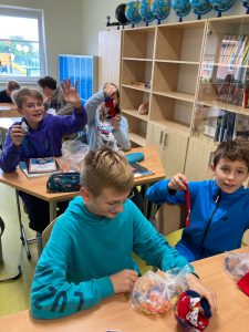 Uczniwie klasy siódmej siedzą przy ławkach w klasie. Na stolikach mają paczki z cukierkamii kolorowe skarpetki.