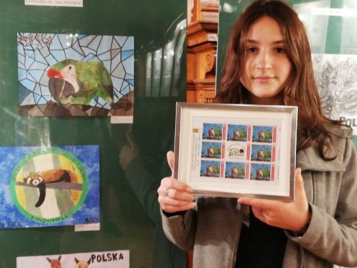 Zdjęcie uczennicy Julii Falkowskiej trzymającej oprawione w ramkę i wydrukowane znaczki jej projektu. Uczennica stoi na tle wystawy na której widać jej pracę.