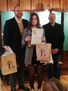 Julia Falkowska pozuje do zdjęcia z dwoma Panami. Uczennica trzyma w dloniach otrzymany dyplom i nagrody w papierowej torbie.