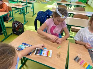Uczniowie podczas tworzenia modeli z plasteliny. Troje dzieci siedzi przy stoliku, naktórym leżą pudełka z kolorową plasteliną. Dziewczynki formują z plasteliny kulki.