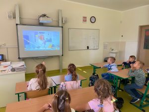 Uczniowie siedzą przy stolikach w sali lekcyjnej i oglądają wyświetlaną na projektorze grafikę.