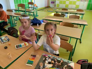 Dwie uczennice siedzą przy wspólnej ławce. Jedna z dziewczynek w yciagniętej dłonitrzyma model wykonany z kulek plasteliny oraz wykałaczek. Druga ma na głowie papierową białą opaskę. Przed dziewczynkami na stolkach leżą przybory - wykałaczki i kolorowe plasteliny.