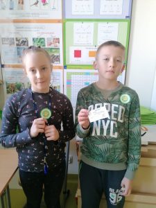 Dwoje uczniów klasy trzeciej pozuje do zdjęcia z odznakami i certyfikatami mistrza tabliczki mnożenia otrzymanymi za poprawne wykonanie działań.