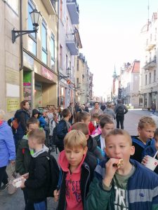 Uczniowie klas II i III idą ulicą szeroką w Toruniu. Część dzieci je kanapki, inne trzymają napoje.