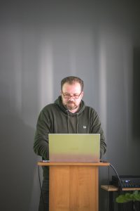 Łukasz Kazek stoi za mównicą, na której stoi otwart laptop.