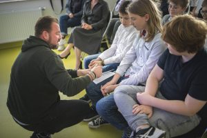 Łukasz Kazek kuca przy uczniach siedzących na krzesłkach w rzędzie. Na kolanie jednego z uczniów nasz gość opiera książkę, którą podpisuje.