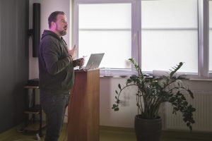 Łukasz Kazek podczas swojej prelekcji. Gość naszej szkoły stoi przy mównicy, w jednej dłoni trzyma okulary wa drugą podnosi w górę na wysokość piersi..