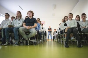 Uczniowie naszej szkoły siedzą na krzesełkach podczas prelekcji Łukasza Kazka. W połowie zdjęcia jest przerwa między siedzącymi uczniami, na końcu której stoi rozmazana postać.