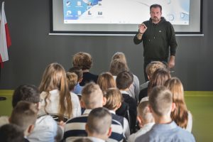 Łukasz Kazek podczas prelekcji na sali teatralnej. Na zdjęciu widać rząd uczniów na przeciwk, której stoi Gość naszej szkoły z podniesioną w górę prawą dłonią.
