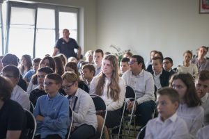 Uczniowie siedzą na krzesełkach na sali teatralnej. Słuchają prelekcji Łukasza Kazka.