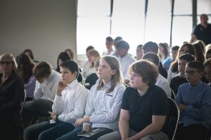 Uczniowie siedzą na krzesełkach na sali teatralnej podczas prelekcji gościa naszej szkoły Łukasza Kazka. Uczniowie zasłuchani patrzą przed siebie.