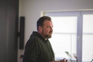 Zdjęcie portretowe Łukasza Kazka wykonane podczas wystąpienia w sali teatralnej. Na zdjęciu uchwycony prawy profil prelegenta.
