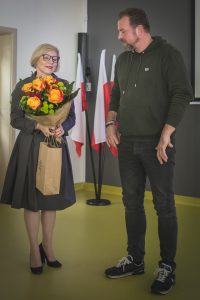 Pani Dyrektor trzyma kwiaty i prezent w papierowej torbie, które za moment przekaże stojącemu obok Łukaszowi Kazkowi.