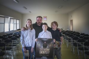 Łukasz Kazek pozuje do zdjęcia z trójką uczniów, którzy podczas prelekcji wygrali nagrody. Nasz gość obejmuje dwoje z tych uczniów ramieniem a przed nimi stoi trzeci chłopiec. Każdy z uczniów w dłoniach trzyma wygraną nagrodę.