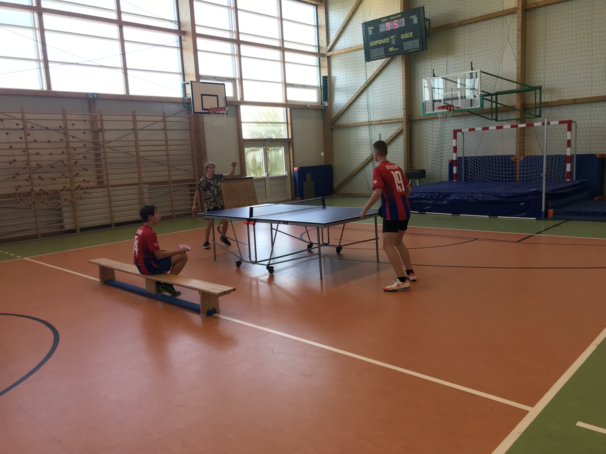 Uczniowie na sali gimnastycznej grają w tenisa stołowego. Dwóch uczniów gra. Trzeci uczeń siedzi na ławeczce obok.