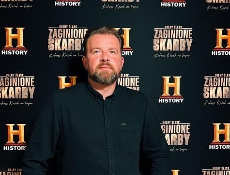 Zdjęcie Łukasza Kazaka stojącego na tle czarnej ściany z logo kanału telewizyjnego HISTORY oraz tytułem programu: ŻAGNIONE SKARBY.