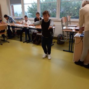 Uczeń klasy czwartej podczas wykonywania skłony. Chłopiec ma wyciagnietew przód dłonie. W tle widać siedzących przy połaczonych stołach innych uczniów.