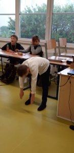 Uczeń klasy czwartej podczas wykonywania skłonu. W tlewidać fragment sali i dwóch chłopców siedzących przy stoliku.