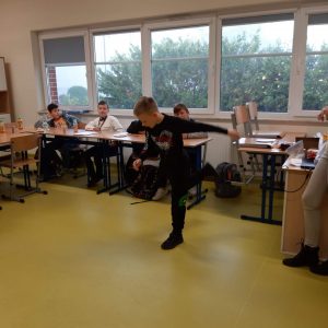 Uczeń klasy czwartej podczas wykonywania wymachów prawą nogą. W tle widać siedzących przy połączonych stołach innych uczniów.