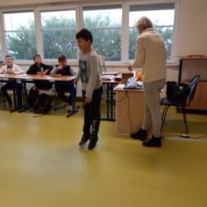 Uczeń klasy czwartej podczas wykonywania podskoku. W tle widać siedzących przy stole pozostałych ucznióworaz odwruconą tyłem nauczycielkę.