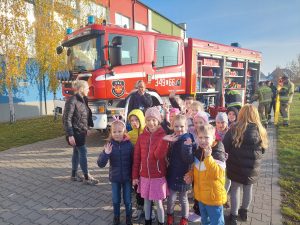 Uczniowie klasy pierwszej stoją przed wozem strażackim. Dzieci machają do zdjęcia. Obok widać Panią Pedagog a z tyłu przy wozie strażackim strażaków.