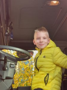Uczeń klasy pierwszej siedzi za kierownicą wozu strażackiego. Chłopiec ma żółtą kurtkę i lekko się uśmiecha.