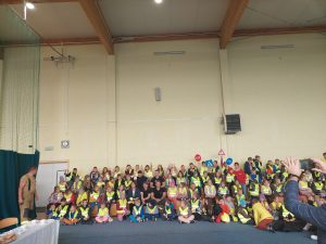 Uczniowie różnysz szkół siedzą na podłodze i ławeczkachw jednej grupie na dużej sali. Dzieci mają założone żółte odblaskowe kamizelki.