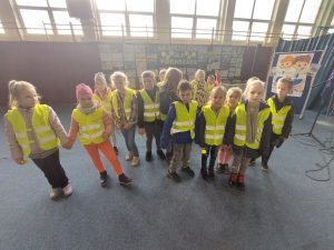 Uczniowie klasy pierwszej w zółtych odblaskowych kamizelkach stoją obok siebie na sali gimnastycznej. W tle widać przygotowanądekorację z napisem "KAMIZELKA DLA PIERWSZAKA".