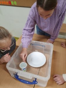 Uczennica pochyla się nad plastikowym pudełkiem, w którym umieszczono biały talerzyk i kubek. Dziewczynka próbuje delikatnie położyc spinacz na tafli wody w kubku. Z boku przyglada się oparta o ławkę jej koleżanka.