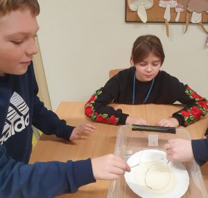 Uczennica siedzi przy stoliku i patrzy przed siebie. Obok stoi uczeń trzymający wykonane z drucika kólko. Pod kółkiem widać biały talerz z płynem.