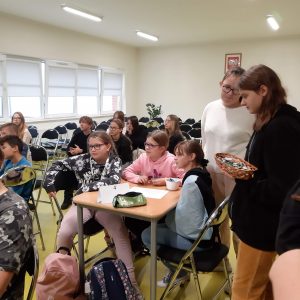 Uczniowie podczas konkursu wiedzy o Janie Pawle II. Przy stoliku widać trzy uczennice klasy piątej, nad którymi stoją ich starsze koleżanki z klasy szóstej. Jedna z nich trzyma koszyczek z cukierkami.