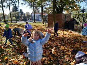 Uczniowie oddziału przedszkolnego chodzą wśród liści na placu przy szkole. Dziewczynka stojąca na pierwszym planie podnosi wysoko w górę ręce.