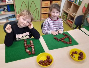 Dwie uczennice oddziału przedszkolnego siedzą przy wspólnej ławce. Przed nimi na zielonych podkładkach leżą ułożone z kasztanów litery A, a przed podkładkami miseczki z kasztanami.