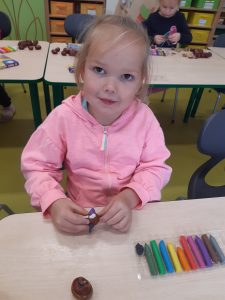 Przy stoliku siedzi uczennica oddziału przedszkolnego. Dziewczynka trzyma w dłoniach kolorową plastelinę, a na stoliku leży plastelina w pudełku i kasztan. W tle widać inne dzieci pracujące przy swoich stolikach.