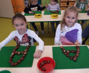 Dwie uczennice oddziału przedszkolnego siedzą przy wspólnej ławce. Przed nimi na zielonych podkładkach leżą ułożone z kasztanów litery A, a pomiędzy miseczka z kasztanami. W tle widać inne dzieci siedzące przy stolikach.