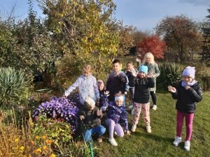 Uczniowie oddziału przedszkolnego stoją wśród zielonych drzew i krzewów. Dzieci machają do zdjęcia. Ustawieni są w jednej grupce. Część z nich kuca.