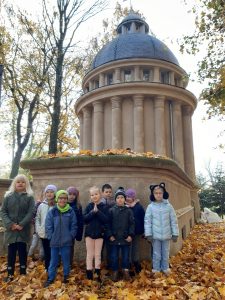 Uczniowie oddziału przedszkolnego stoją przy ścianie kapliczki. Na zdjęciu widać górującą nad dziećmi kapliczkę zwieńczoną okrągłą, niebieską kopułą.