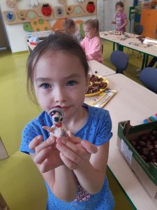 Uczennica oddziału przedszkolnego trzyma w dłoniach wykonaną przez siebie figurkę z plasteliny. W tle widać pracujące inne dzieci.