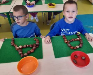 Dwóch uczniów oddziału przedszkolnego siedzą przy wspólnej ławce. Przed nimi na zielonych podkładkach leżą ułożone z kasztanów litery A, a pomiędzy miseczka z kasztanami. W tle widać inne dzieci siedzące przy stolikach.