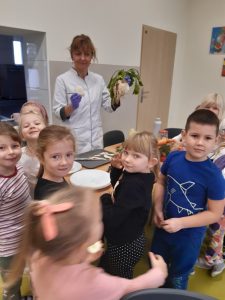 Uczniowie oddziału przedszkolnego gromadzą się za stołem na szkolnej stołówce. Po drugiej stronie stołu stoi ubrana w biały kitel Pani nauczycielka trzymająca w dłoni rzepę z zieloną natką.